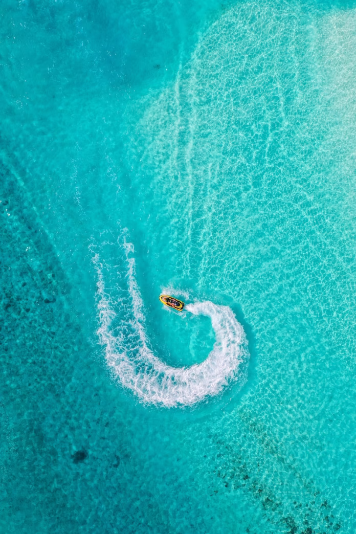 Aerial view of a tourist riding a jet ski through the crystal-clear turquoise waters of Fulhadhoo Island in Baa Atoll Biosphere Reserve, Maldives, with pristine white sand beaches and coral reefs visible below
