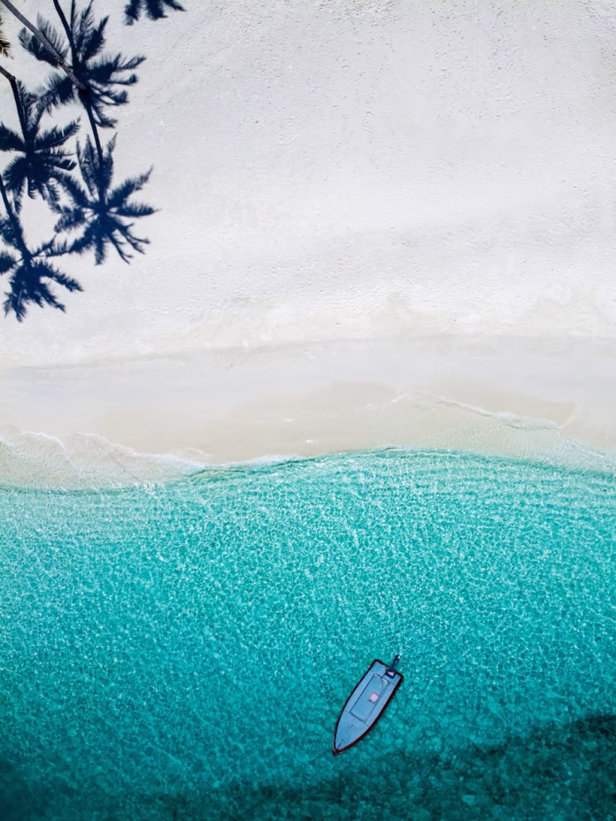 Aerial view of Feridhoo Island's curved white sand beach in Ari Atoll, Maldives, with coconut palms lining the shore and turquoise lagoon fading to deep blue