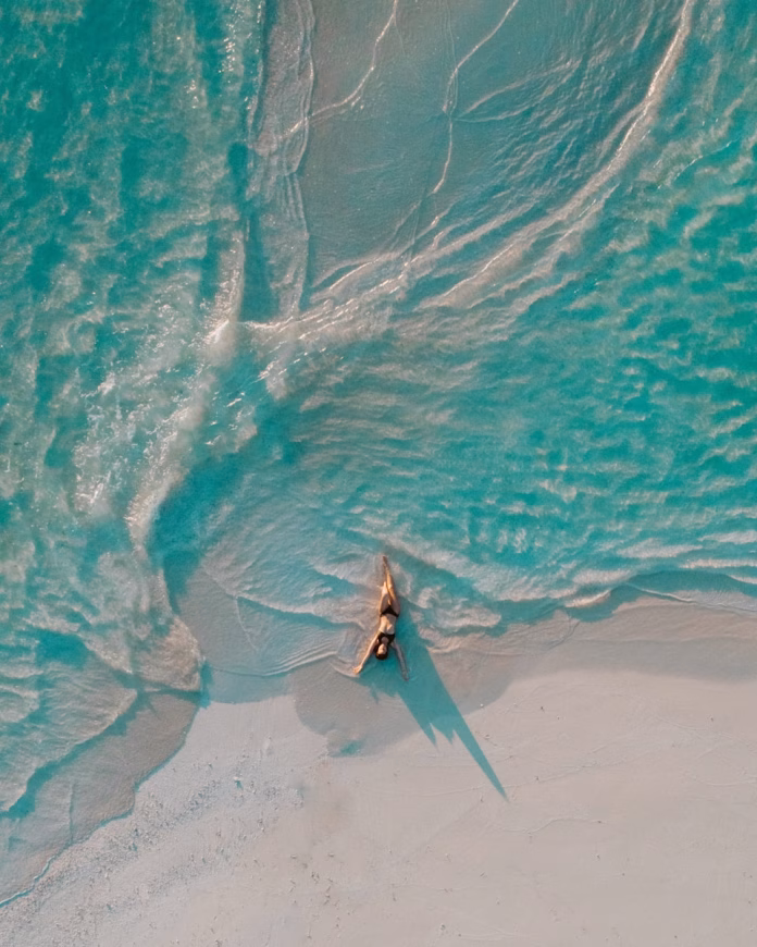 Aerial view of a guest relaxing on Gulhi Island's empty white sand beach in South Malé Atoll, with turquoise shallows fading to deep blue in the Maldives