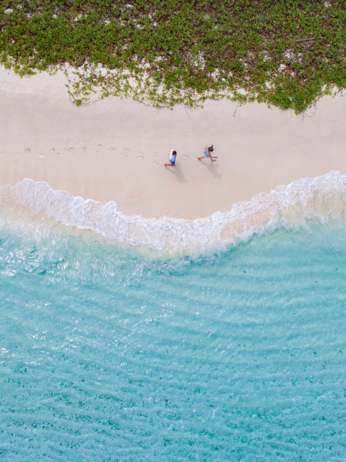 Aerial view of a couple walking hand-in-hand along Gulhi Island's pristine white sand beach in South Malé Atoll, Maldives, with turquoise lagoon and coral reefs visible in the crystal-clear waters