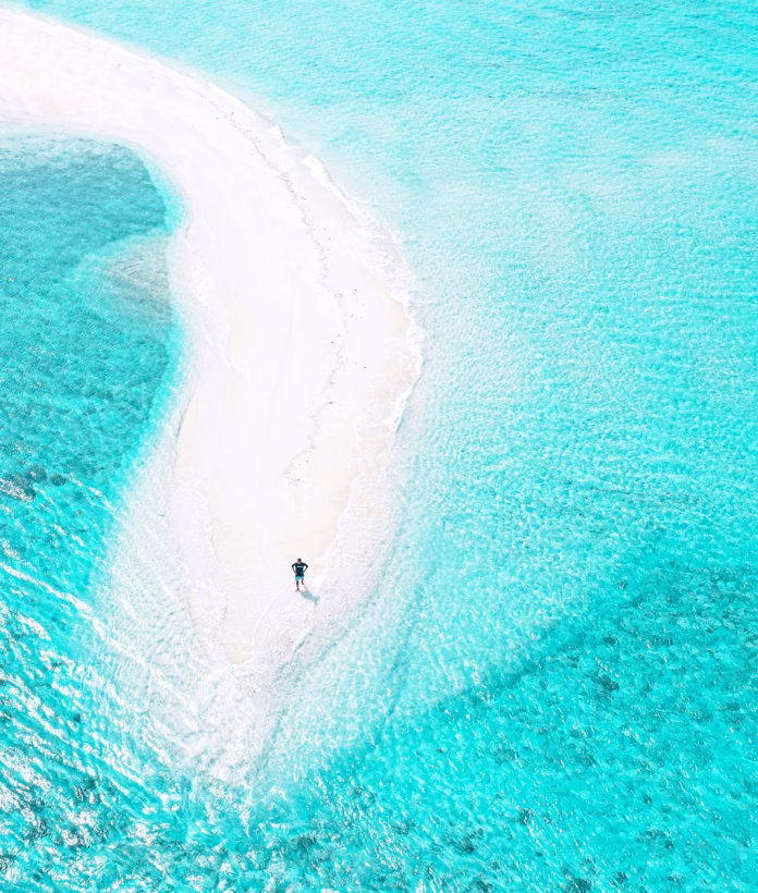 Solitary traveler walking on Omadhoo's endless sandbank at low tide, surrounded by mirror-like turquoise waters in Baa Atoll, Maldives